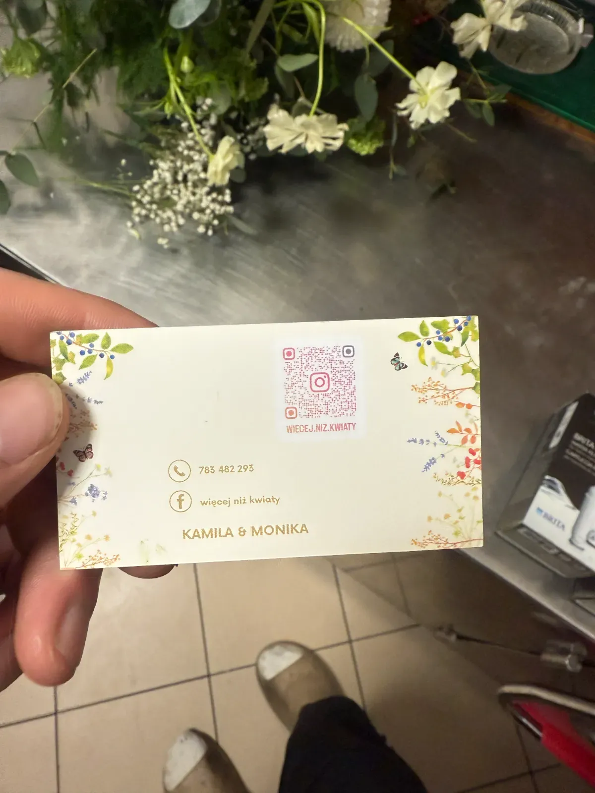Wizytówka kwiaciarni trzymana w dłoni, w tle bukiet na blacie