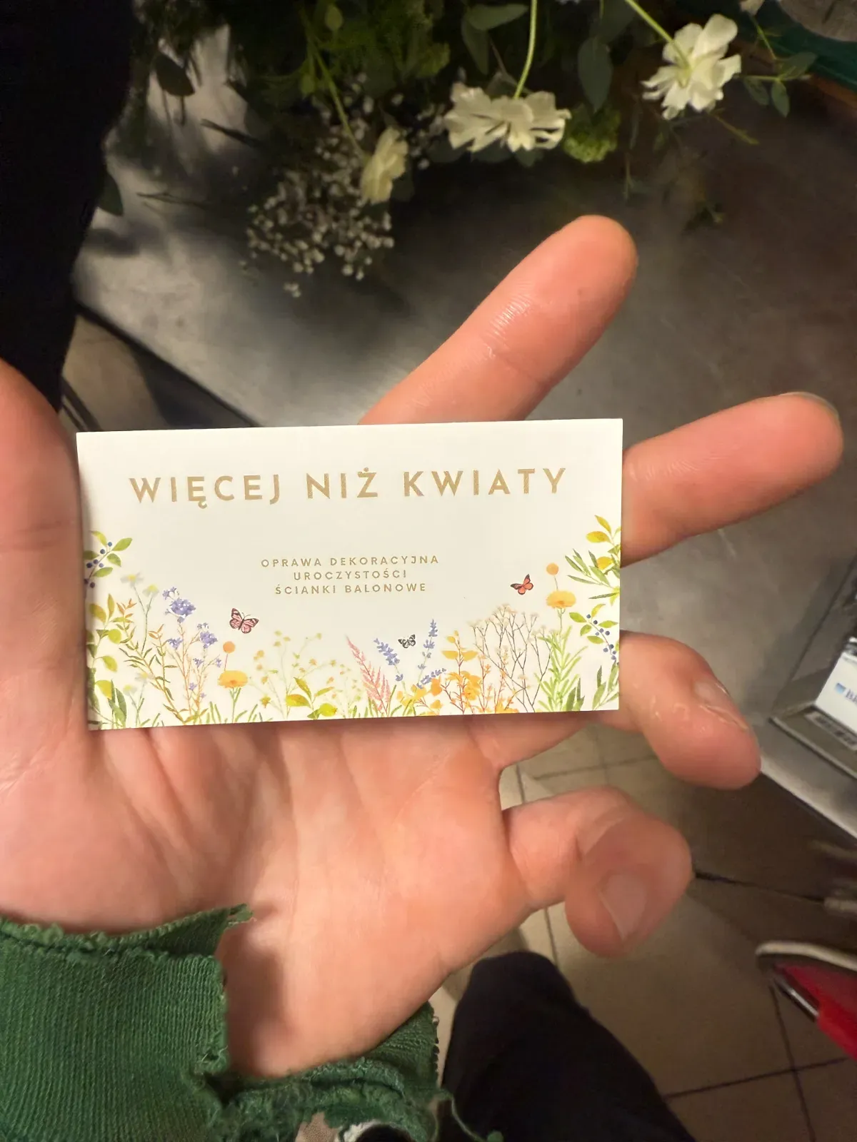 Wizytówka firmy dekoratorskiej w dłoni, za nią bukiet kwiatów